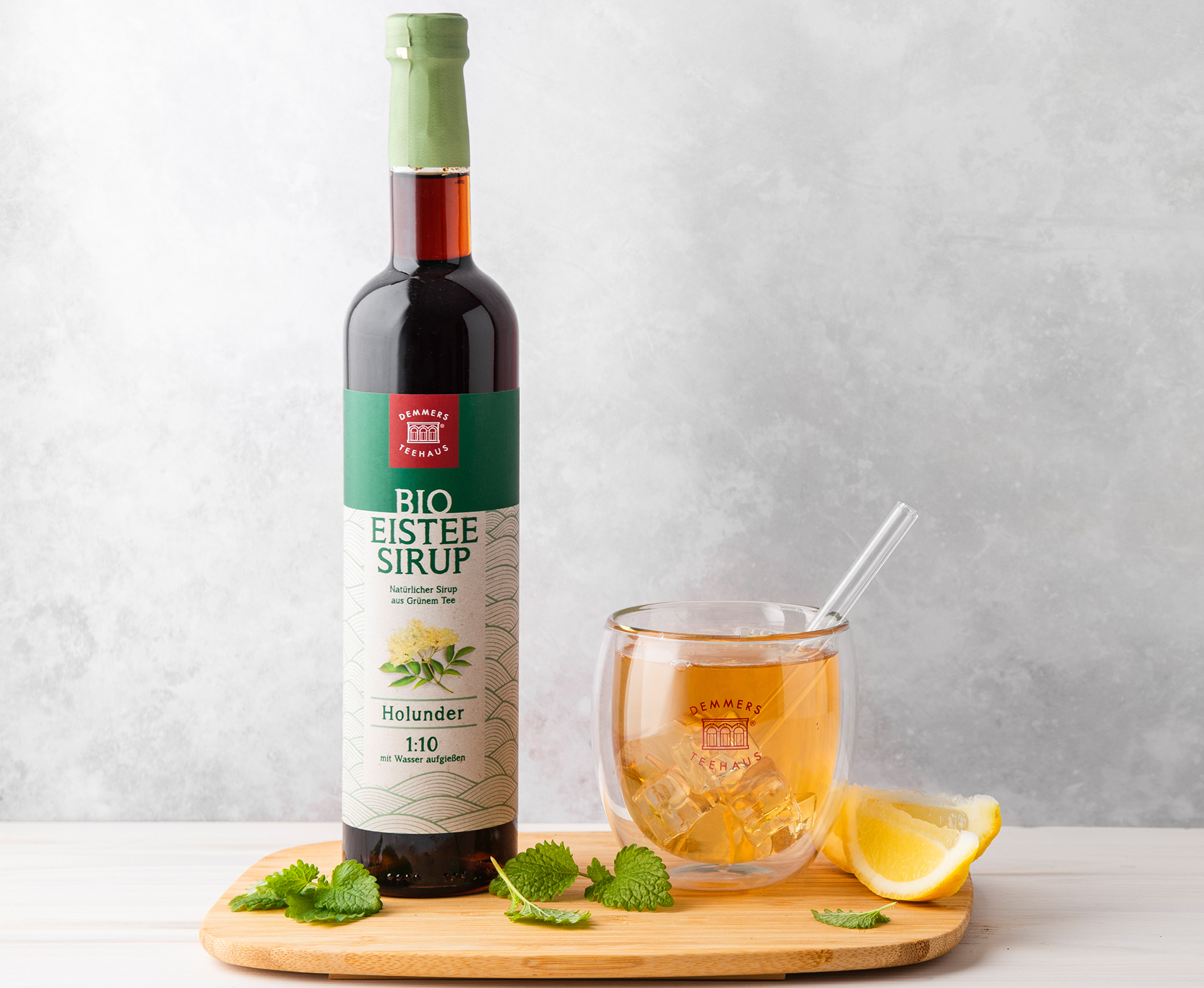 Demmers Teehaus BIO Eistee-Sirup Grüner Tee Holunder Flasche neben Glas mit kaltem Eistee und Limetten auf Holztablett